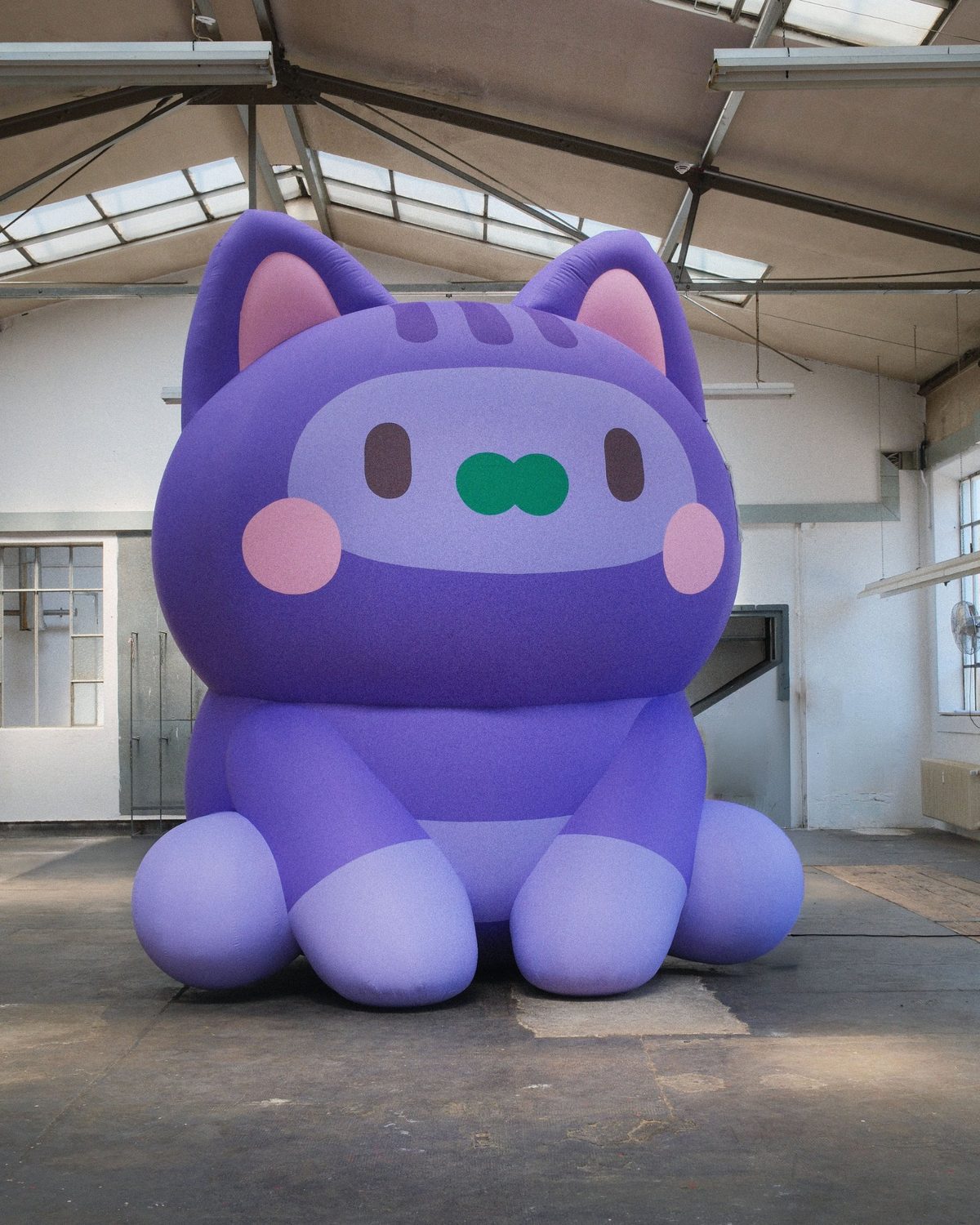 The giant purple inflatable cat with pink cheeks and a green nose sitting in an empty hall