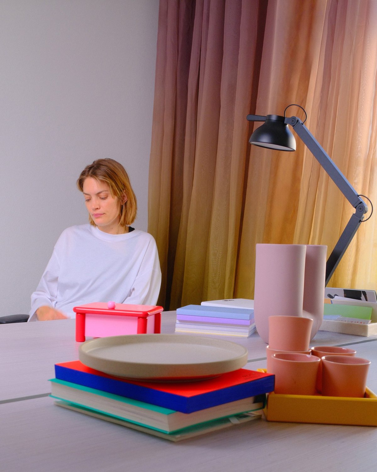 A woman at a table behind The Dot, a red and pink jewellery box on round legs, with colourful books and pink ceramics