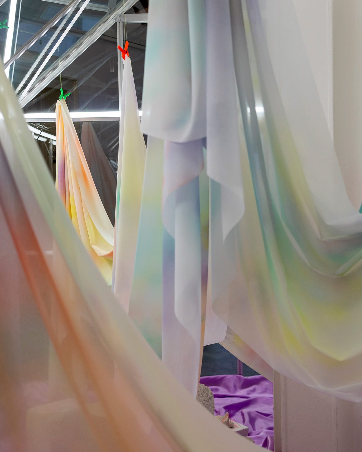 Looking through draped, airbrushed silk towards pieces hanging from neon clips inside the greenhouse