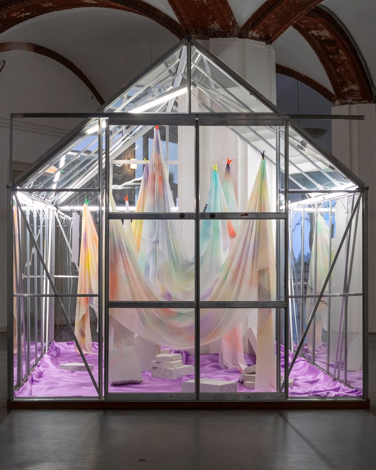 A glass greenhouse filled with pastel, hand-painted silk hanging from colourful clips over a lilac floor